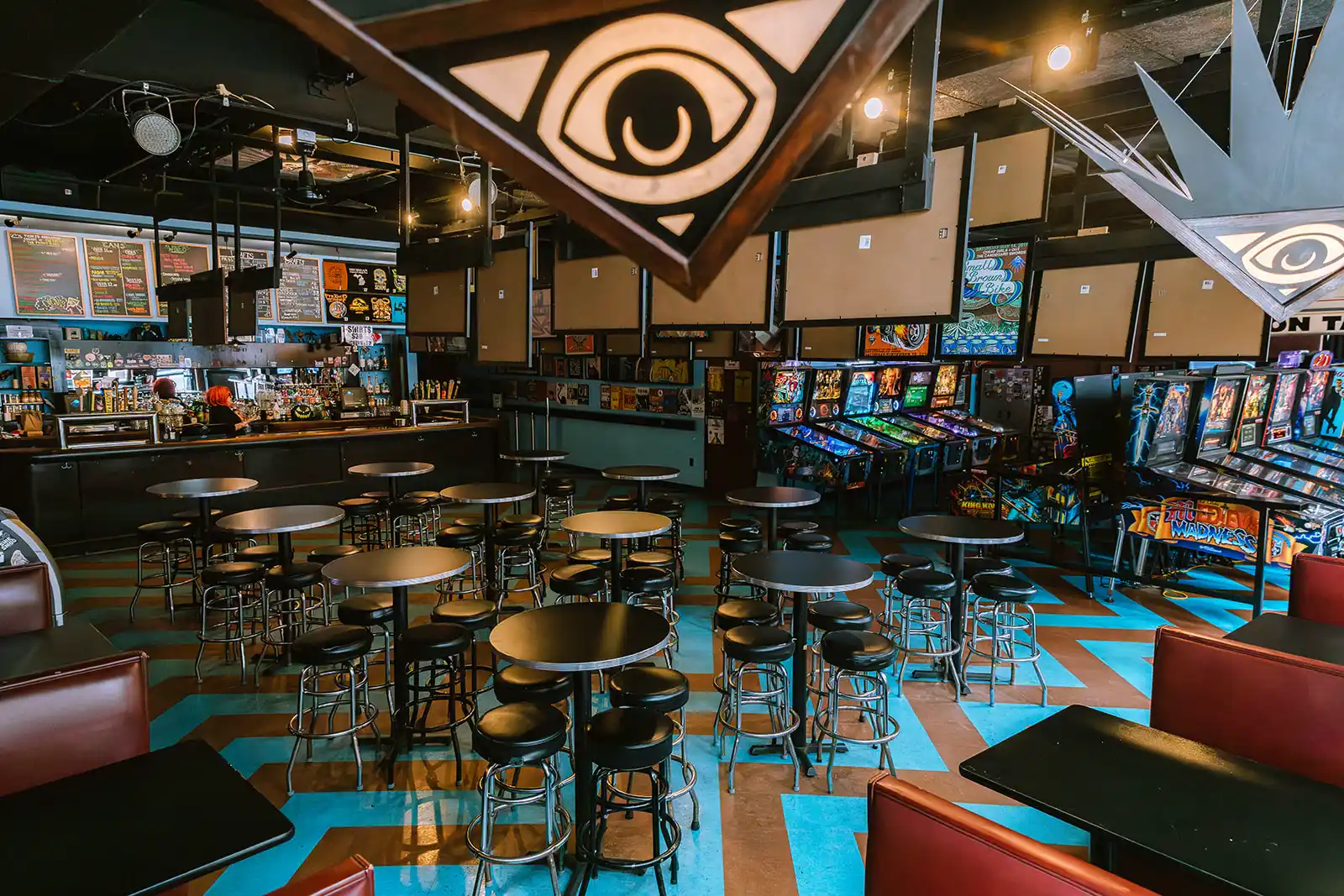 wide view of bar area with hightop tables and bar stools, full bar to left, pinball machines to right, and logo eye hanging from ceiling