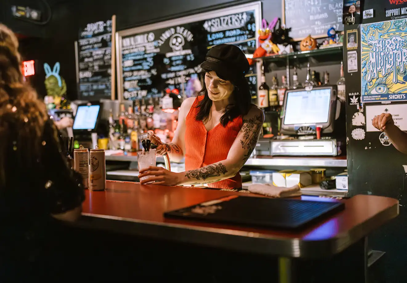 smiling bartender in orange tank top and black hat pouring a drink into a glass with ice