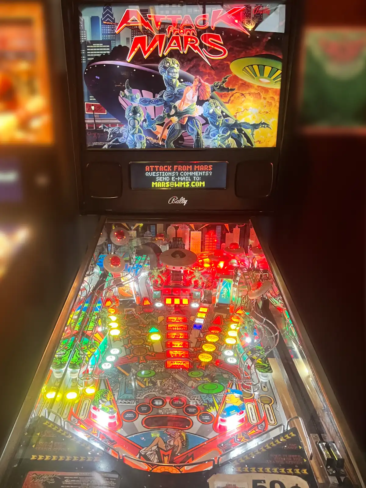 A brightly lit "Attack from Mars" pinball machine with a colorful playfield full of red, yellow, and green lights. The backglass shows attacking aliens and a flying saucer over a cityscape.