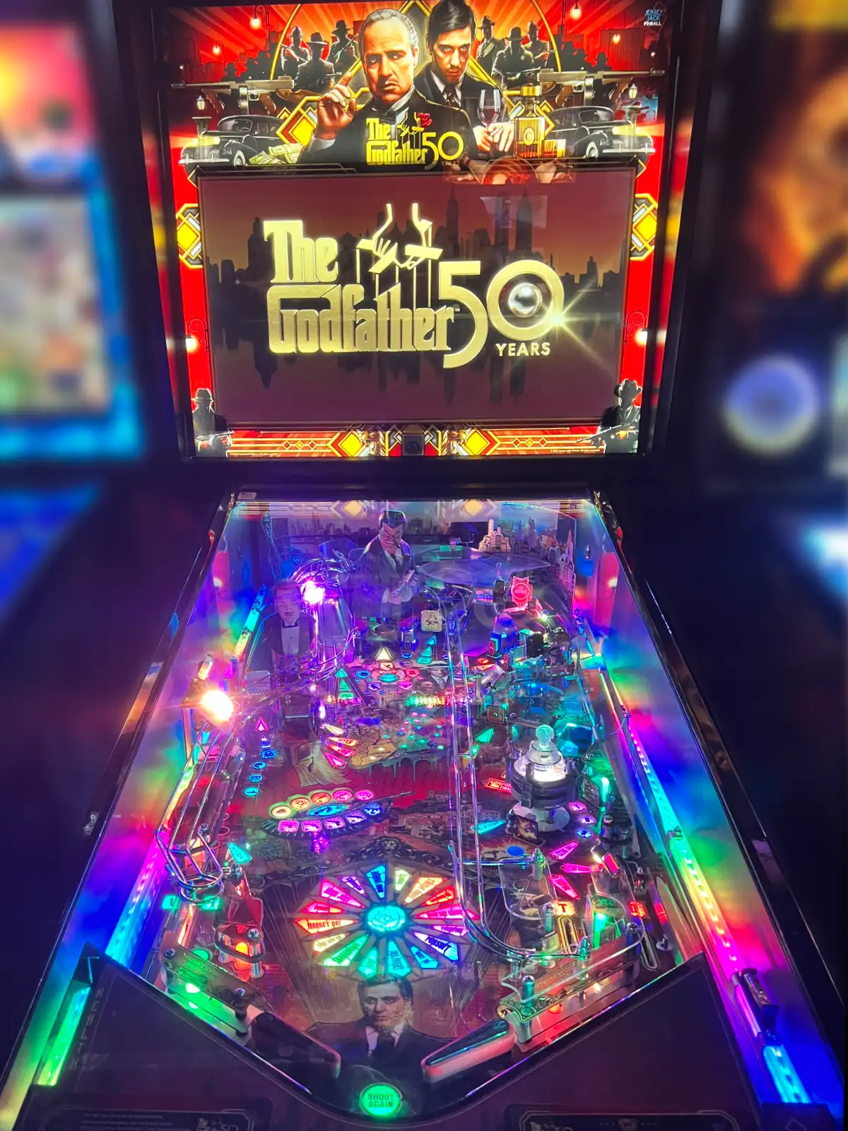 A brightly lit "The Godfather 50 Years" pinball machine glows with vibrant pink, green, and blue LED lights across the complex playfield.