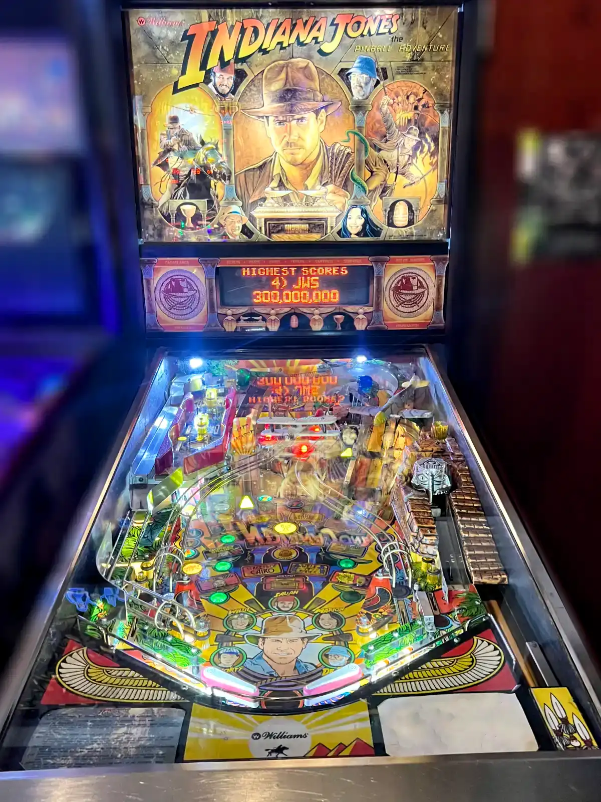 A brightly illuminated, vertical shot of the Indiana Jones pinball machine showing the colorful playfield, movie graphics, and a high score of 300 million.