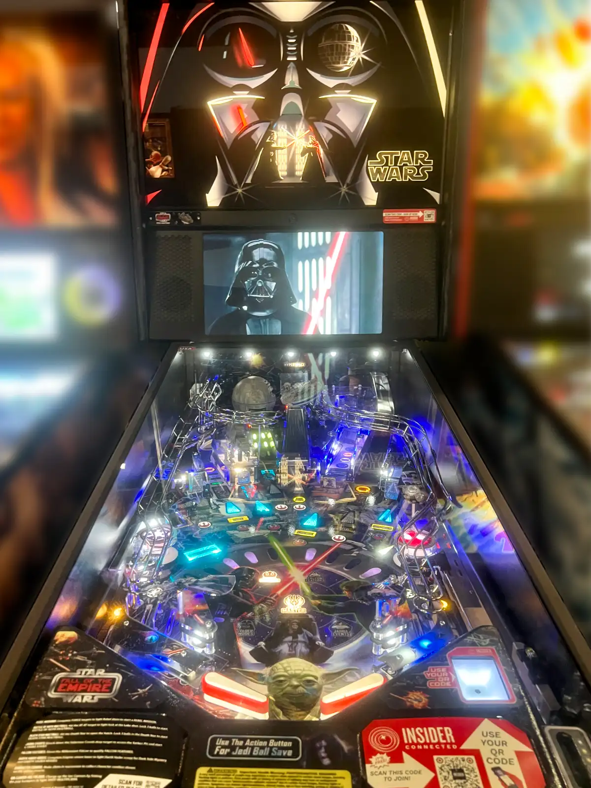 A Star Wars: Fall of the Empire pinball machine, brightly illuminated with blue lights, featuring Darth Vader on the back panel and a Yoda figure.