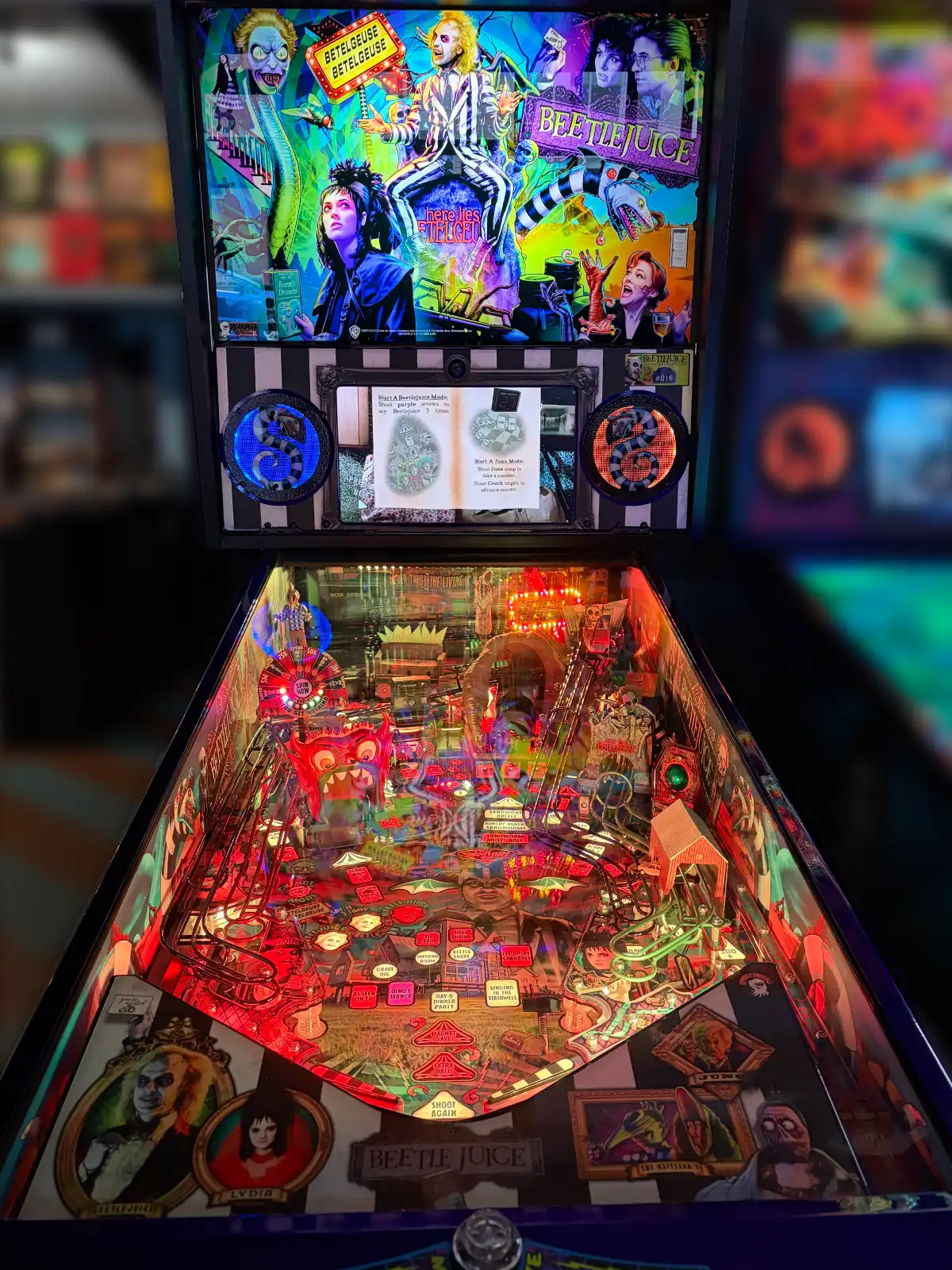 A vibrant Beetlejuice pinball machine glows intensely red and purple, displaying the film's characters on the backbox and the complex, detailed playfield.