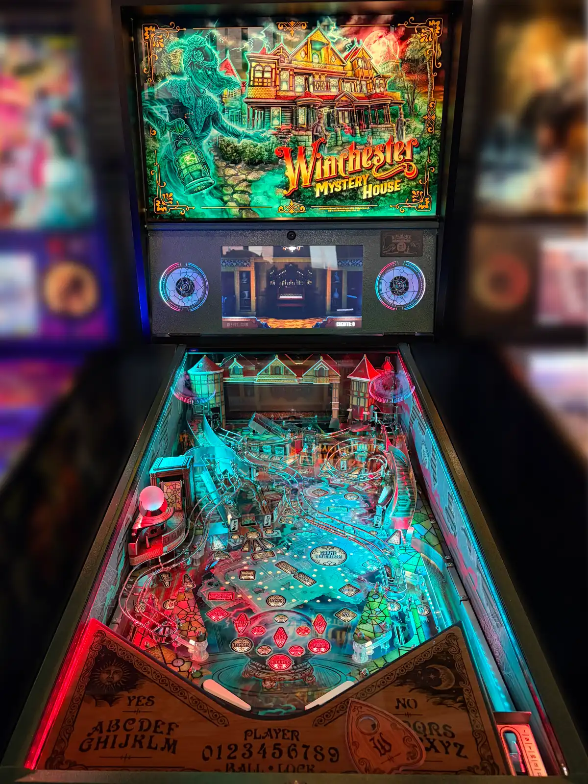A Winchester Mystery House themed pinball machine shows the detailed playfield and back-glass art, lit brightly in teal and red, including a Ouija board motif.