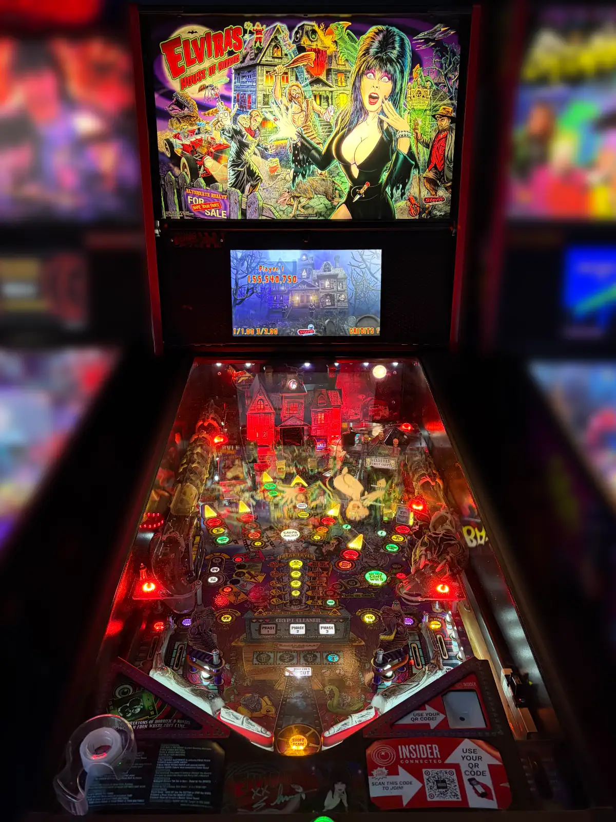 An "Elvira's House of Horrors" pinball machine illuminated in red light, showing the playfield, backglass art, and the high score display.
