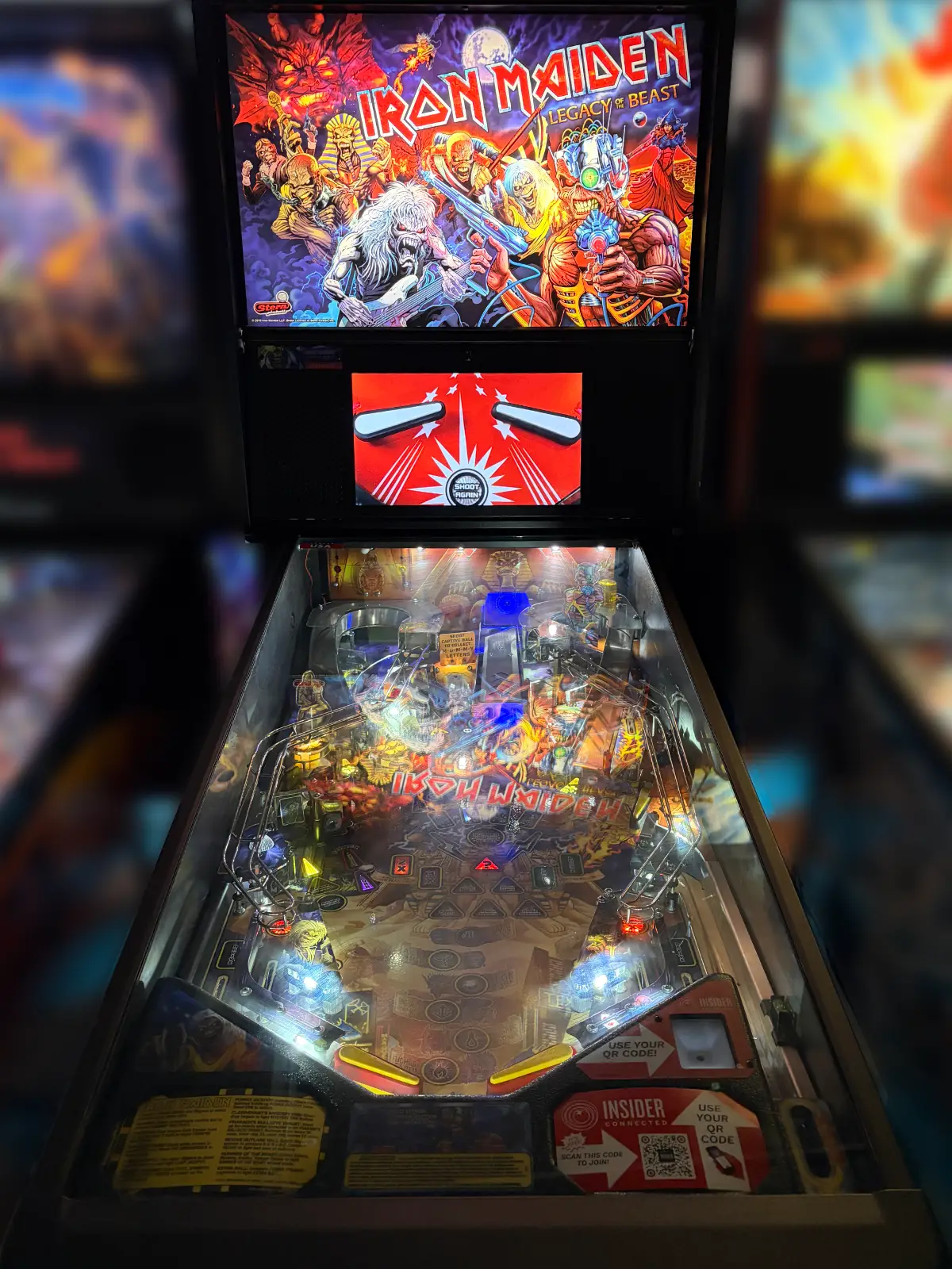 A Stern Iron Maiden: Legacy of the Beast pinball machine is brightly lit in a dark room. The machine's backglass displays aggressive band artwork, while the detailed playfield shines beneath the glass.
