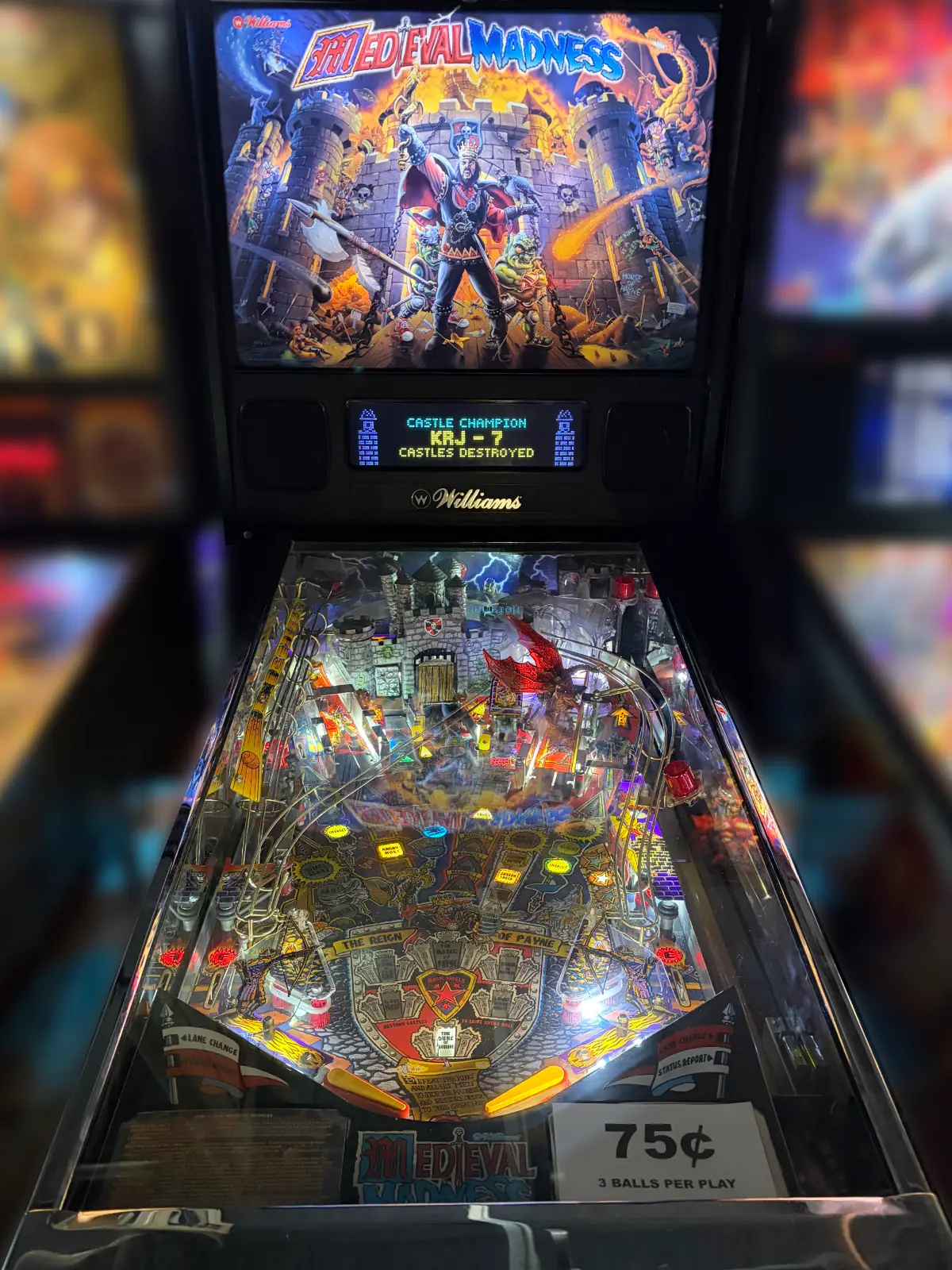 A Williams "Medieval Madness" pinball machine illuminated in a dim venue. The digital display reads "Castle Champion KRJ-7." A sign shows "75¢ 3 Balls per Play."