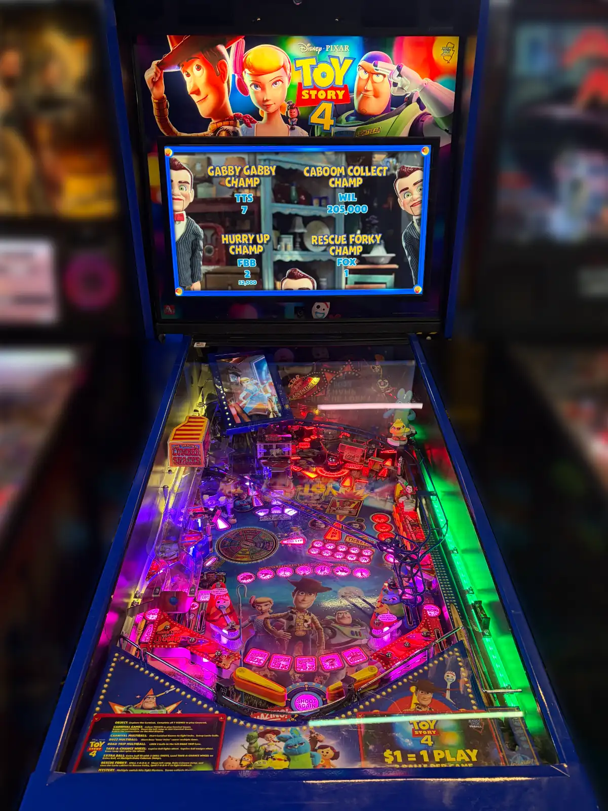 A close-up view of a Toy Story 4 pinball machine with bright pink and blue lights illuminating the busy playfield. The upper screen displays the movie logo and high score champion titles.